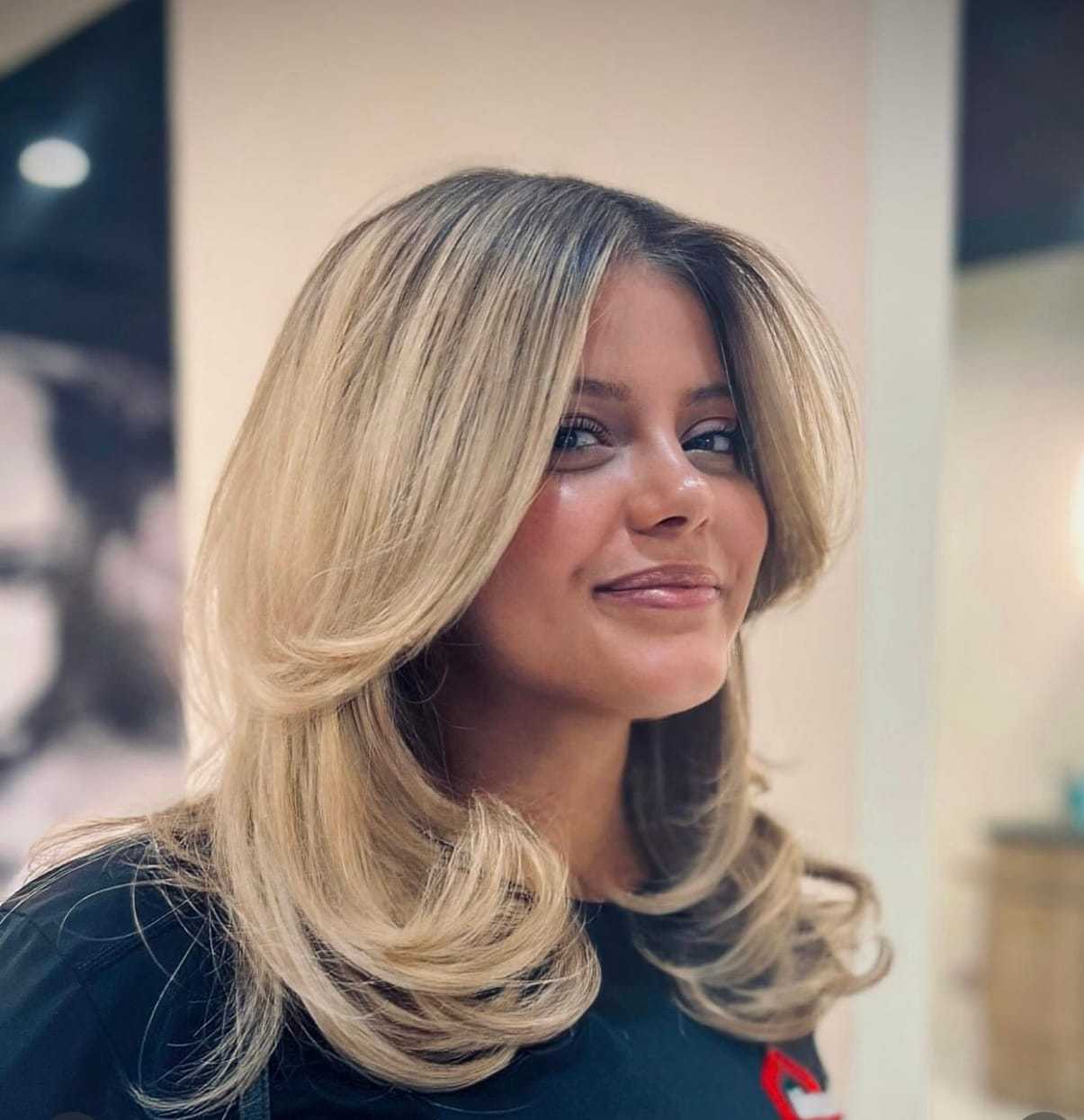 Woman with blonde, wavy hair smiling at the camera in a brightly lit setting.